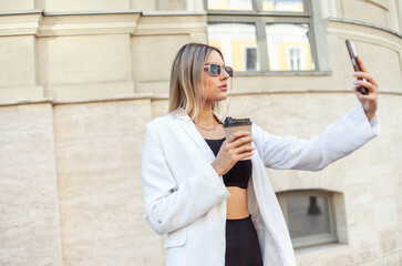 Fototapeta premium Young fashion blonde woman in trendy outfit drinks coffee on the go and makes selfie on phone in city