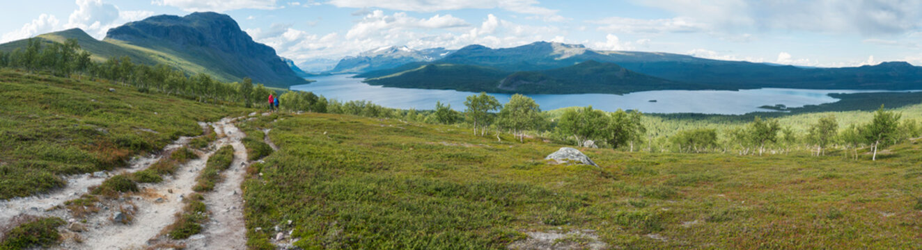 Panoramic landscape with beautiful river Lulealven, snow capped mountain, birch trees and two hikers couple at Kungsleden hiking trail near Saltoluokta, Sweden, Lapland wild nature. Summer blue sky