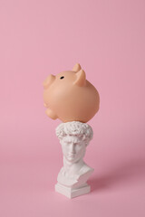 Antique bust of David with piggy bank on pink background. Conceptual pop. Minimal still life.