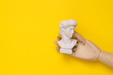 Wooden hand holding David bust on yellow background. Minimal still life