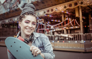 lifestyle portrait extraordinary hipster woman with african braids dressed in a denim jacket with a skateboard in hand at amusement park