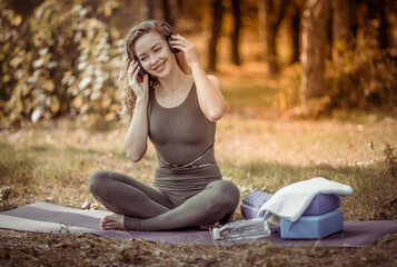 Attractive active yogi woman with curly hair listens to relaxing music with headphones while sitting on a mat in the forest