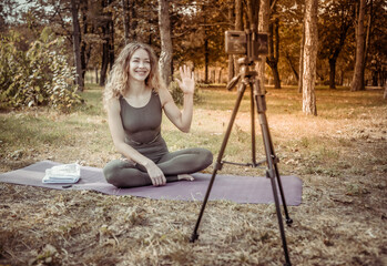 Young woman yoga teacher leads online lesson in forest. Sport woman broadcasts to camera of smartphone standing on a tripod. Blogging, vlog. Healthy lifestyle