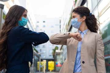 Two business woman with masks bump elbows to avoid coronavirus outdoors