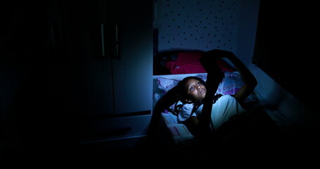 Fototapeta na wymiar Teen black girl using cellphone device at night in bed