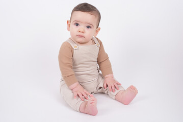 Adorable baby boy wearing beige overalls sitting on white background looking at camera and smiling. 