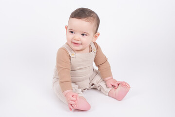 Adorable baby boy wearing beige overalls sitting on white background looking aside and smiling. 