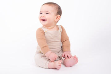 Adorable seven month old baby boy wearing beige overalls sitting on the floor over white background...