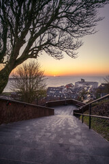 Sunrise through under the tree and above small port city of Helgoland, Germany. Sunrise colors in the sky. Sun disc rising on the distant horizon. Stair leading down to the city.