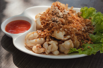 Garlic fried squid in a plate garnished with greens and served with spicy chili sauce is set on the dining table with natural daylight,  Seafood menu that is popular with both children and adults.