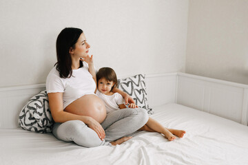 Pregnant expecting mother relaxing with little girl daughter in white bedroom interior at home. Waiting child, preparation for childbirth. Happy family concept.