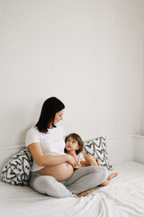 Pregnant expecting mother relaxing with little girl daughter in white bedroom interior at home. Waiting child, preparation for childbirth. Motherhood concept.