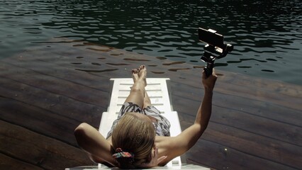 Woman shooting on handheld film gimbal stabilization for smartphone. Girl rest, relax, lie sunbed on pier in sunglasses, make selfie. Lady blogger broadcast video blogging vlogging. Take photo video. © ivandanru