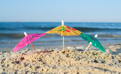 Fototapeta na wymiar Three small paper cocktail umbrellas stand in sand on sandy beach close-up. Small paper umbrellas on sandy shore near sea and sea waves on sunny summer day. Leisure, Vacation, Travel, Tourism Concept