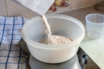 Wholemeal bread flour is being tipped from a white paper bag into a plastic bowl on weighing scales. Selective focus.