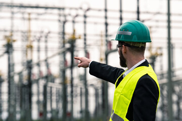 Electric engineer wearing helmet suit and safety vest at bad weather pointing at substation 