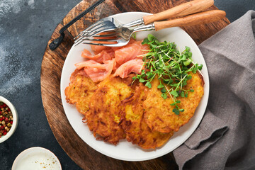 Potato pancakes. Fried homemade potato pancakes or latkes with cream, green onions, microgreens, red salmon and sauce in rustic plate on old black table background. Rustic style. Top view.