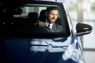 Smiling bearded man in stylish suit sitting inside new blue car during visit of modern showroom. Successful businessman examining various auto before making purchase.