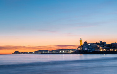 Obraz premium Fotografia de larga exposición de Sitges desde la playa con la iglesia iluminada al fondo en un atardecer, anochecer con cielo de colores azul, naranja y amarillo y unas ligeras nubes en el horizonte