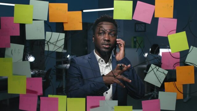 African-American businessman talk on cellphone looking at glassboard with notes in dark office