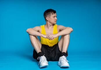 Fototapeta na wymiar thin athlete in a yellow t-shirt and black shorts on a blue background. funny shakes muscles, goes in for sports, fitness