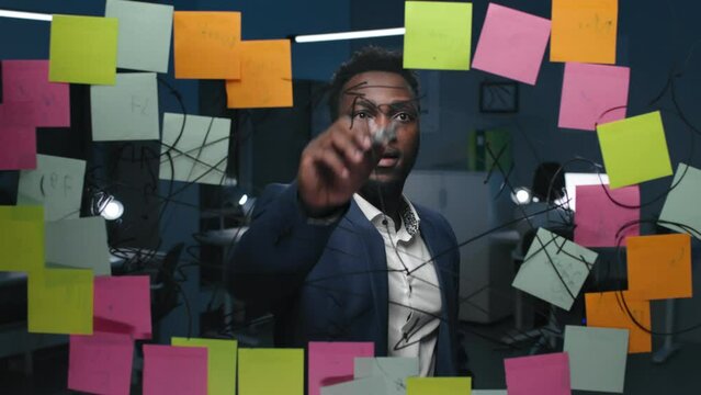 African-American businessman write mindmap on glassboard in dark office. 