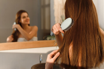 Smiling pretty brunette in a towel combs her long shiny silky hair with a comb in the morning after cosmetic procedures, on the background of the bathroom.