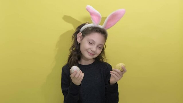 4k. Happy easter. Beautiful cute little girl in Easter bunny ears holding two painted easter eggs on a yellow background. Easter activities for children. Hello spring concept
