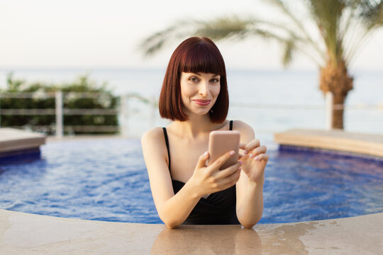 Gorgeous young smiling woman in black swimsuit, using smartphone at outdoor swimming pool at beautiful sea ocean resort. Woman sharing vacation photos with her friends