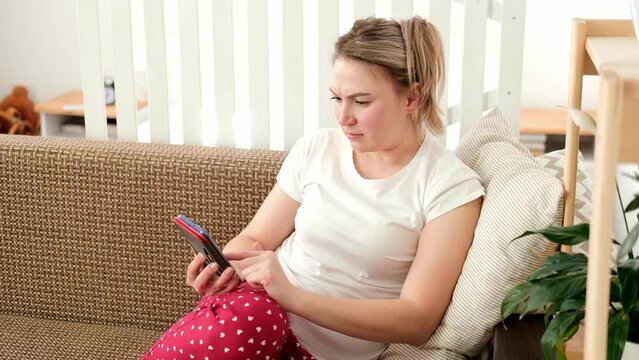 Caucasian woman is sitting at home on couch, looking at smartphone screen and frowning. Reads bad news, gets upset. Concept of mental burnout and doom scrolling.