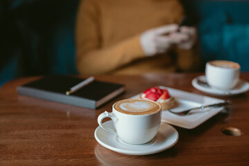 Cup of coffee, delicious cute strawberry tart, black notebook on wooden table, man checking his mobile phone on background. Man having a break in a coffee shop.
