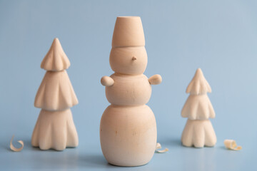 Handmade wooden carved snowman stands on blue background. Blurred Christmas trees with few shavings in the background. Winter decorations theme.