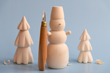 Handmade wooden carved snowman with carving knife stands on blue background. Blurred Christmas trees with few shavings in the background. Winter decorations theme.