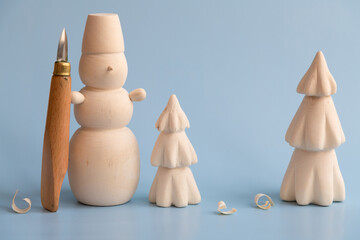 Handmade wooden carved snowman with carving knife stands next to Christmas tree with few shavings on blue background. Copy space for your text. Winter decorations theme.