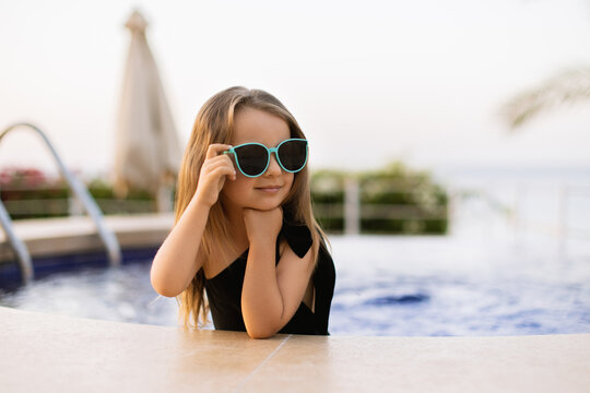 Adorable little girl at swimming pool in a black swimsuit and blue sunglasses having fun during summer vacation. Young girl posing near swimming pool touching the glasses with her hand.