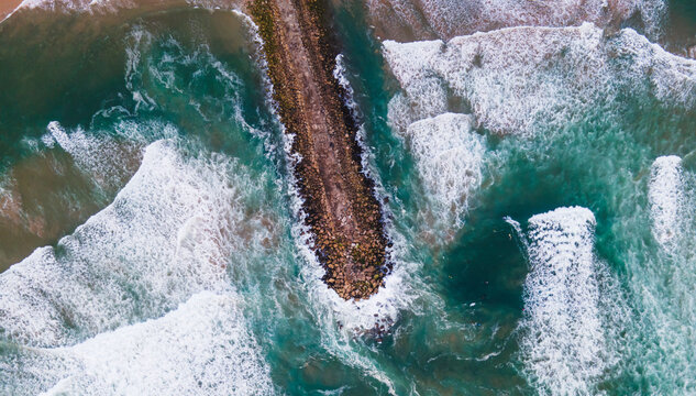 Aerial view of a majestic breakwater with rough rolling water waves rolling on the beach at Costa da Caparica, Setubal, Portugal.