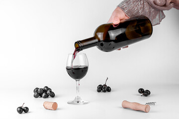 Human Hand pouring red wine from bottle into glass, grape bunches and corkscrew on grey background. Wine degustation concept.