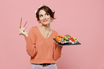 Young minded pensive thoughtful fun woman in sweater hold in hand makizushi sushi roll served on black plate traditional japanese food look aside chopsticks isolated on plain pastel pink background
