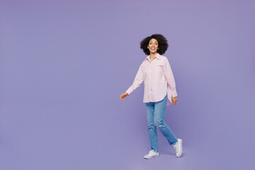 Full body smiling happy fun cheerful young woman of African American ethnicity 20s wear pink striped shirt walking going look camera isolated on plain pastel light purple background studio portrait.