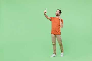 Full body young man 20s in casual orange t-shirt doing selfie shot on mobile cell phone post photo on social network show victory gesture isolated on plain pastel light green color background studio