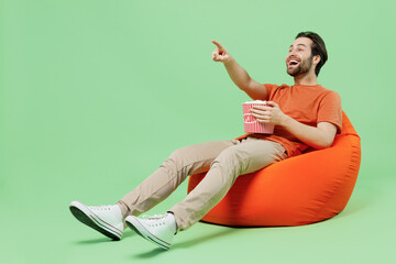 Full body young man 20s wear casual orange t-shirt sit in bag chair hold takeaway bucket of popcorn...