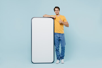 Full body young man wear yellow t-shirt stand near big mobile cell phone with blank screen workspace area show thumb up isolated on plain pastel light blue background studio. People lifestyle concept.
