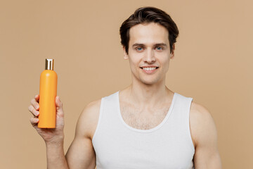 Attractive cool fun young man 20s perfect skin in undershirt hold bottle of shampoo balsam isolated...