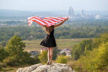 Young pretty american woman with long hair holding waving on wind USA flag on her sholders standing outdoors enjoying warm summer day.