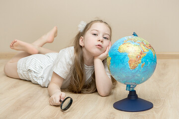 pensive little girl lies on the floor next to a globe, holds a magnifying glass in one hand, put the other under her head and argues. The concept of a smart child, education, plans for the future