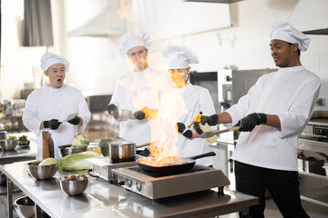 Multiracial team of professional cooks in uniform preparing meals for a restaurant in the kitchen. Latin guy burning pan, european cooks making sauce and asian chef managing the process. Teamwork and