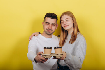 Two young beautiful people girl and guy standing on a yellow background. They drink coffee