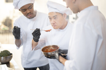 Three chef cooks with different ethnicities tasting sauce with a spoon while cooking in the kitchen. Teamwork and cooking delicious food at restaurant