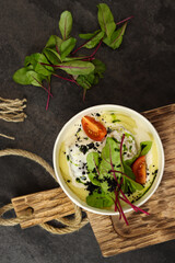 Mashed potatoes with fish in cream sauce, tomatoes and beetroot leaves in paper container on wood board isolated on dark background. Healthy food delivery service and daily ration concept.