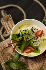 Mashed potatoes with fish in cream sauce, tomatoes and beetroot leaves in paper container on wood board isolated on dark background. Healthy food delivery service and daily ration concept.